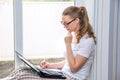 Home quarantine. Remote work at home. Caucasian woman sitting at window with a computer.Counts on calculator and writes in a Royalty Free Stock Photo