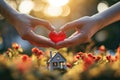 Home sweet home concept. Dream housing. Real estate. Two hands holding a red heart over a miniature house outdoors on a sunny