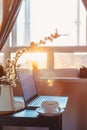 Home work place with laptop, cup of hot drink and blooming brunch in vase on coffee table near window on sunset or sunrise. Royalty Free Stock Photo