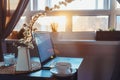 Home work place with laptop, cup of hot drink and blooming brunch in vase on coffee table near window on sunset or sunrise. Royalty Free Stock Photo