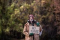 Homeless beggar man standing outdoors in park, holding bag and cardboard sign.