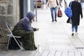 Homeless beggar sat on busy street wearing a hoodie with cup for change in the UK with shoppers in the background