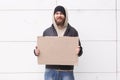 A homeless European-looking man stands on the street near a wall with a cardboard sign and smiles. Outdoor. Royalty Free Stock Photo