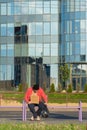 A homeless guy sits on the bench with a cardboard and an inscription: need money. In the background is a business center