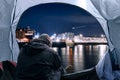 Homeless man hand on his head in despair wearing dark cloths in a tent by a river. Illuminated city in the background out of focus Royalty Free Stock Photo