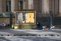 Homeless man sleeps on the street outside a stone building beside a information poster