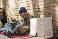 Homeless Man Using Laptop Sitting By The Brick Wall In The Street.