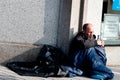 A homeless person siting down holds a cup begging for money while covered in a sleeping bag