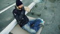 Homeless young man in dirty clothes drink alcohol sitting near shopping cart on the street at cold winter day