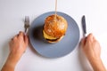 Homemade cheeseburger on a gray plate and a person with cutlery in his hands on a white background. Hands in the frame Royalty Free Stock Photo