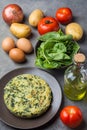 Homemade delicious frittata with spinach on plate. Recipe ingredients potatoes eggs olive oil in bottle tomatoes on dark concrete