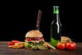 Homemade hamburger with french fries and bottle of beer on wooden table. In the burger stuck a knife. Fastfood on dark background Royalty Free Stock Photo