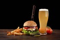 Homemade hamburger with french fries and glass of beer on wooden table. In the burger stuck a knife. Fastfood on dark background Royalty Free Stock Photo