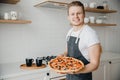 Homemade Italian pizza, handsome young male chef in dark apron holds baking sheet in bright kitchen. Cooking video blog Royalty Free Stock Photo