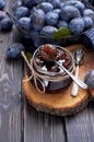 Homemade plum jam in a glass jar and fresh blue plums in a bowl on a dark rustic wooden background with copy space top view.