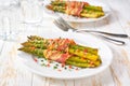 Homemade roasted bundles of green asparagus wrapped in bacon on a ceramic plate on a white wooden table, close up Royalty Free Stock Photo