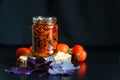 Homemade sun-dried tomato slices in glass jar in olive oil, basil, oregano spices, red pomodoro on dark background.