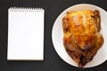 Homemade traditional rotisserie chicken on a white plate, blank notepad on a black surface, top view. Flat lay, overhead, from