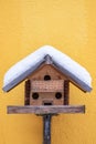 Homemade wooden bird`s feeder on the yellow wall in winter, under snow