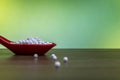 Homeopathic pills in red spoon and some granules scattered on wood surface with green yellow background Royalty Free Stock Photo