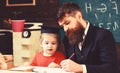 Homeschooling concept. Teacher in formal wear and pupil in mortarboard in classroom, chalkboard on background Royalty Free Stock Photo