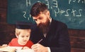 Homeschooling concept. Teacher in formal wear and pupil in mortarboard in classroom, chalkboard on background. Father Royalty Free Stock Photo