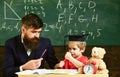 Hometask concept. Teacher in formal wear and pupil in mortarboard in classroom, chalkboard on background. Father Royalty Free Stock Photo