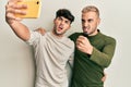 Homosexual gay couple standing together taking a selfie photo with smartphone annoyed and frustrated shouting with anger, yelling