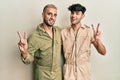 Homosexual gay couple standing together wearing casual jumpsuit smiling looking to the camera showing fingers doing victory sign Royalty Free Stock Photo