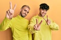 Homosexual gay couple standing together wearing yellow clothes smiling looking to the camera showing fingers doing victory sign Royalty Free Stock Photo