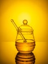 Honey in a glass jar with a stick on a yellow background. A jar of honey is reflected off the surface of the table.