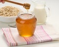 Honey in a glass transparent jar and a wooden stick on a white table, behind a decanter with milk and oatmeal in a plate
