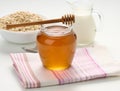 Honey in a glass transparent jar and a wooden stick on a white table, behind a decanter with milk and oatmeal in a plate