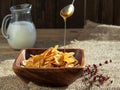 Honey from a spoon is poured into corn cereal. Jug of milk , corn cereals in a wooden plate on dark wooden table. Closeup, Royalty Free Stock Photo