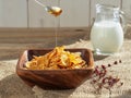 Honey from a spoon is poured into corn cereal. Jug of milk , corn cereals in a wooden plate on dark wooden table. Closeup, Royalty Free Stock Photo