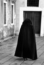 hooded anonymous person walking wearing an old black tabard as a cloak on the narrow alley of the city