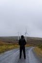 A hooded figure standing on a dirt track. Looking at wind turbines. On a bleak mountain track. Wales. UK