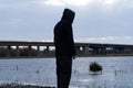 A hooded figure, standing by a lake in the countryside in winter