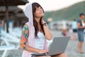 A hooded happy woman with a tattoo on her arm sits on a chaise longue and works on a laptop. Side view. Resort. The concept of Royalty Free Stock Photo