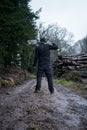 A hooded man, back to camera on a muddy path holding a spade, next to a log pile on a grey moody day in the countryside Royalty Free Stock Photo