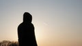 A hooded man standing, back to camera. Watching a plane with a contrail in the sky at sunset