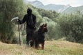 Hooded Person with Dog, Mysterious cloaked figure stands in a natural setting, holding a scythe