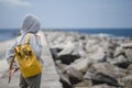 A hooded woman puts on a backpack while standing on an ocean pier. Back view. Royalty Free Stock Photo