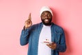 Hopeful Black guy celebrating birthday, making wish with fingers crossed, holding bday cake with candle, standing