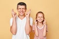 Hopeful happy father and daughter wearing casual t-shirts standing isolated over beige background closed eyes crossed fingers