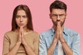 Hopeful woman and man with serious expressions keep hands in praying gesture, have gloomy look, believe in good fortune