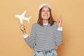 Hopeful woman wearing striped shirt and baseball cap holding white paper plane crossing fingers making wish to travel abroad