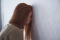 Hopeless and depressed young redhead woman leans her forehead on wall at home