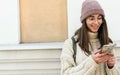 Horizontal image of attractive young woman standing outdoors against building`s wall, browsing on Internet on smart phone. Pretty Royalty Free Stock Photo