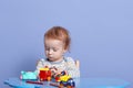 Horizontal image of fair haired sweet child sitting at table, playing with several toys, having fun alone, being focused on game,